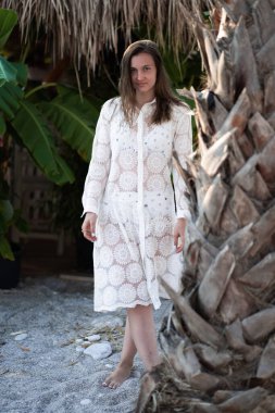 Kumsalda poz veren beyaz plaj elbiseli güzel esmer bir kız. Palm 'ın yanında duran kız. Modaya uygun. Manzaralı. Yaz