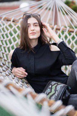 Koyu renk gömlekli, kot pantolonlu ve güneş gözlüklü mutlu kadın gün batımında hamakta dinleniyor. Rahatlatıcı bir kavram. Güzel kız..