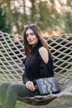 Koyu renk gömlekli, kot pantolonlu ve güneş gözlüklü mutlu kadın gün batımında hamakta dinleniyor. Rahatlatıcı bir kavram. Güzel kız..