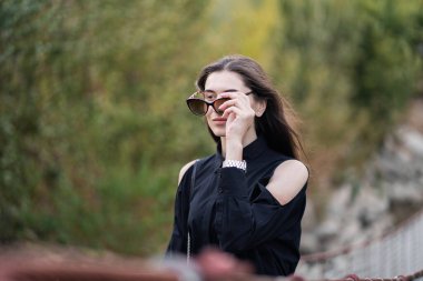 Esmer saçlı, koyu renk elbiseli ve güneş gözlüklü güzel bir kadın. Moda sokak fotoğrafçılığı. Moda modeli, doğa arka planında poz veriyor. Güneş gözlüğü dükkanı