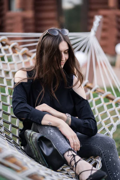 Mulher feliz em camisa escura, jeans e óculos de sol relaxando na rede na floresta ao pôr do sol. conceito relaxante. Menina bonita . — Fotografia de Stock