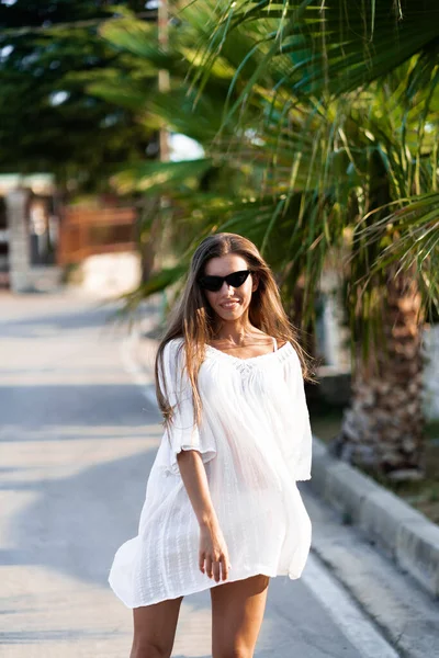 Beyaz elbiseli ve güneş gözlüklü genç ve güzel bir kadın yolda palmiyelerle dönüyor. Tatil boyunca keyif, rahatlık ve özgürlük kavramı. Kız dinlenmenin tadını çıkarıyor. Moda. — Stok fotoğraf