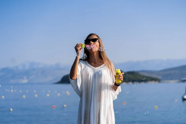 Beyaz elbiseli ve güneş gözlüklü genç bir kadın iskelede sabun köpüğü üflüyor. Tatil boyunca keyif, rahatlık ve özgürlük kavramı. Kız dinleniyor.. — Stok fotoğraf