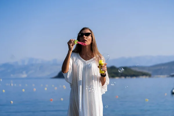 Beyaz elbiseli ve güneş gözlüklü genç bir kadın iskelede sabun köpüğü üflüyor. Tatil boyunca keyif, rahatlık ve özgürlük kavramı. Kız dinleniyor.. — Stok fotoğraf