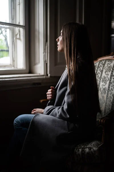 Mujer joven relajándose en casa en silla cómoda. Chica relajante y esperando a alguien. Mujer joven en casa sentada en una silla moderna cerca de la ventana relajándose en la sala de estar y mirando a la ventana . — Foto de Stock