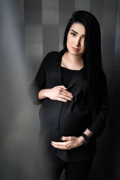 Femme enceinte en vêtements noirs tient la main sur le ventre sur un fond lumineux. Grossesse, maternité, concept de préparation et d'attente - gros plan de la femme enceinte heureuse avec un gros ventre à la fenêtre . — Photo