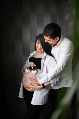 Güzel hamile bir kadın ve bebek ayakkabısı tutan ve göbeğine sarılan yakışıklı kocasının görüntüsü. Hamilelik, annelik, hazırlık ve beklenti konsepti. Dikey