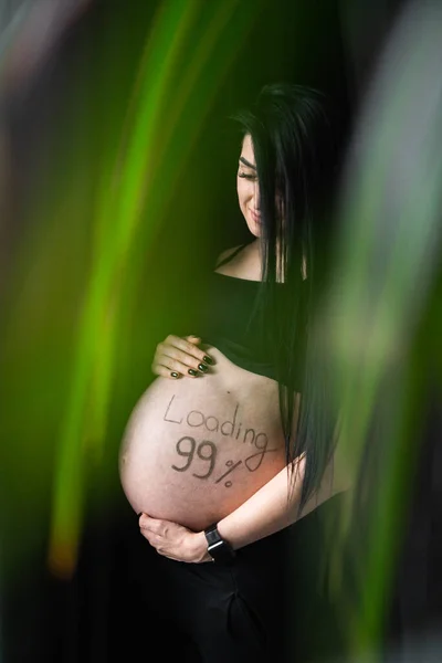 Mujer embarazada en ropa negra toma de la mano en el vientre con la carga de inscripción 99. El embarazo, la maternidad, la preparación y el concepto de expectativa - primer plano de la mujer embarazada feliz con vientre grande. . —  Fotos de Stock