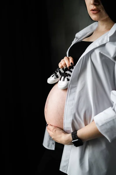 Obrázek krásné těhotné ženy držící dětské boty a objímající bříško. Těhotenství, mateřství, příprava a koncepce očekávání. Svisle — Stock fotografie