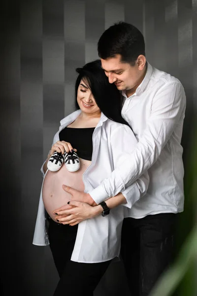 Εικόνα της όμορφης εγκύου και του όμορφου συζύγου της να κρατούν παιδικά παπούτσια και να αγκαλιάζουν την κοιλιά. Εγκυμοσύνη, μητρότητα, προετοιμασία και προσδοκία. Κάθετη — Φωτογραφία Αρχείου