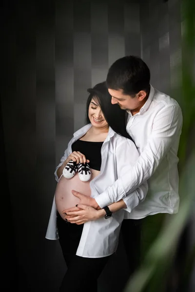 Image d'une belle femme enceinte et de son beau mari tenant des chaussures de bébé et embrassant le ventre. Grossesse, maternité, concept de préparation et attente. Vertical — Photo