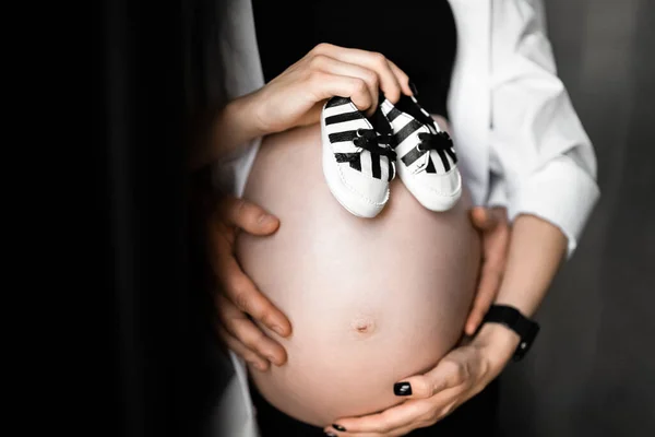 Güzel hamile bir kadın ve bebek ayakkabısı tutan ve göbeğine sarılan yakışıklı kocasının görüntüsü. Hamilelik, annelik, hazırlık ve beklenti konsepti — Stok fotoğraf