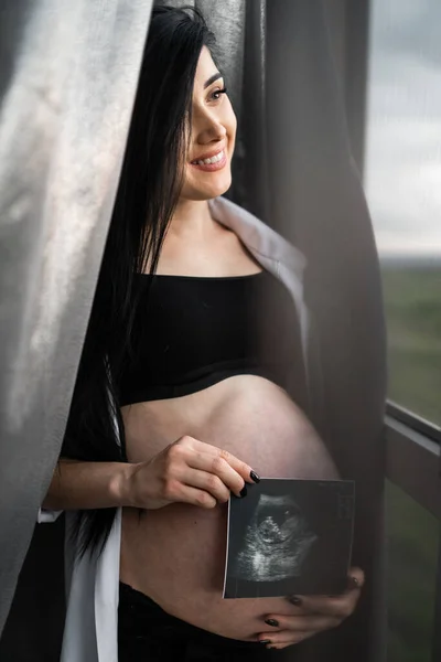 Mujer embarazada con ropa negra sostiene la ecografía en el vientre sobre un fondo oscuro. El embarazo, la maternidad, la preparación y el concepto de expectativa - el primer plano de la mujer embarazada feliz con el vientre grande en la ventana . —  Fotos de Stock