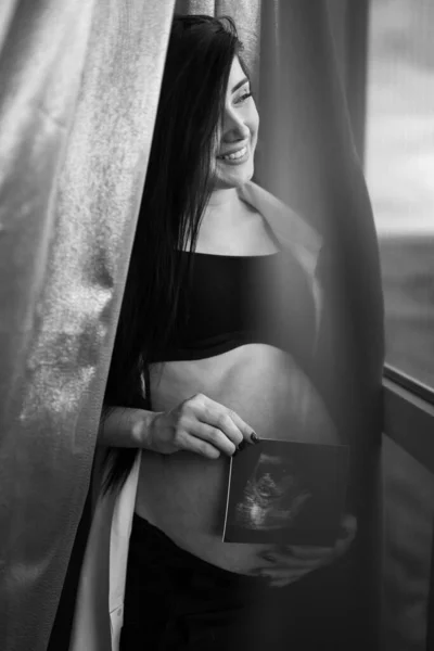 Mujer embarazada con ropa negra sostiene la ecografía en el vientre sobre un fondo oscuro. El embarazo, la maternidad, la preparación y el concepto de expectativa - el primer plano de la mujer embarazada feliz con el vientre grande en la ventana . —  Fotos de Stock