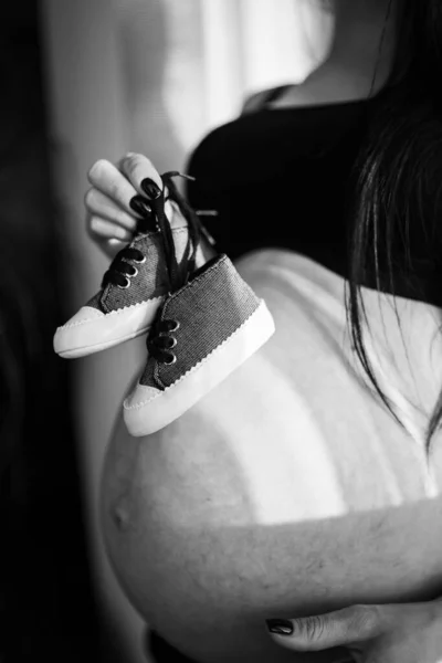 Imagen de una hermosa mujer embarazada sosteniendo zapatos de bebé y abrazando la barriga. Concepto de embarazo, maternidad, preparación y expectativa. Vertical. Primer plano. —  Fotos de Stock