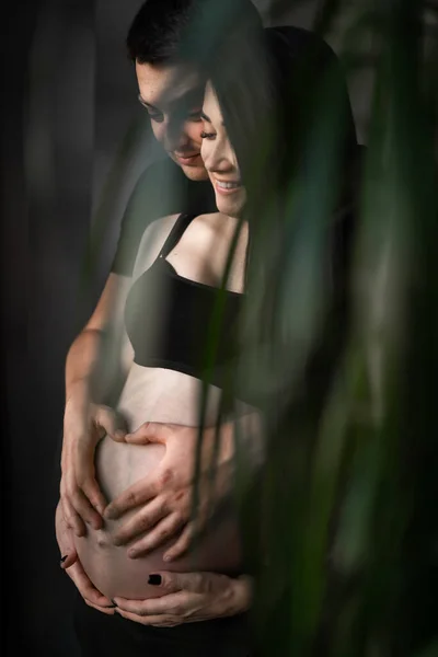 Pareja romántica esperando un bebé, tomados de la mano y tocando la frente mientras están de pie juntos en el vientre contra la ventana en casa, espacio libre. Concepto de embarazo, maternidad, preparación y expectativa . —  Fotos de Stock