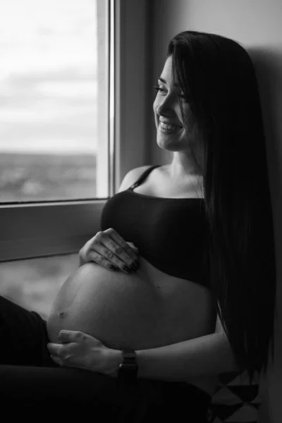 Femme enceinte en vêtements noirs tient la main sur le ventre sur un fond sombre. Grossesse, maternité, concept de préparation et d'attente - gros plan de la femme enceinte heureuse avec un gros ventre à la fenêtre . — Photo