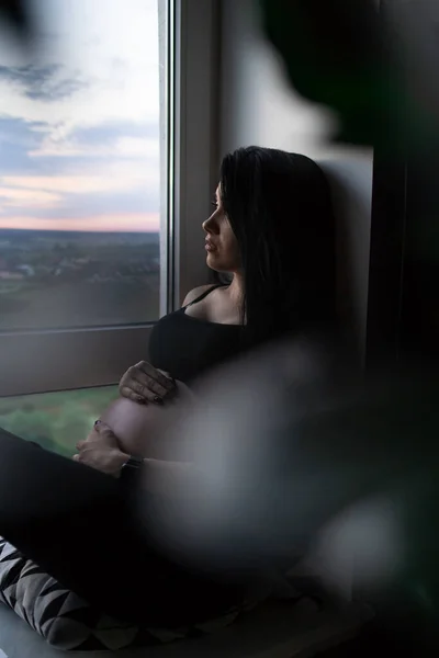Femme enceinte en vêtements noirs tient la main sur le ventre sur un fond sombre. Grossesse, maternité, concept de préparation et d'attente - gros plan de la femme enceinte heureuse avec un gros ventre à la fenêtre . — Photo
