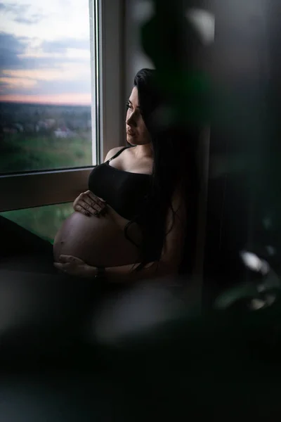 Femme enceinte en vêtements noirs tient la main sur le ventre sur un fond sombre. Grossesse, maternité, concept de préparation et d'attente - gros plan de la femme enceinte heureuse avec un gros ventre à la fenêtre . — Photo