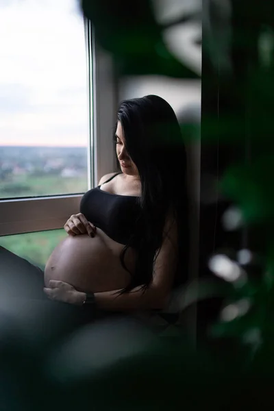 Femme enceinte en vêtements noirs tient la main sur le ventre sur un fond sombre. Grossesse, maternité, concept de préparation et d'attente - gros plan de la femme enceinte heureuse avec un gros ventre à la fenêtre . — Photo