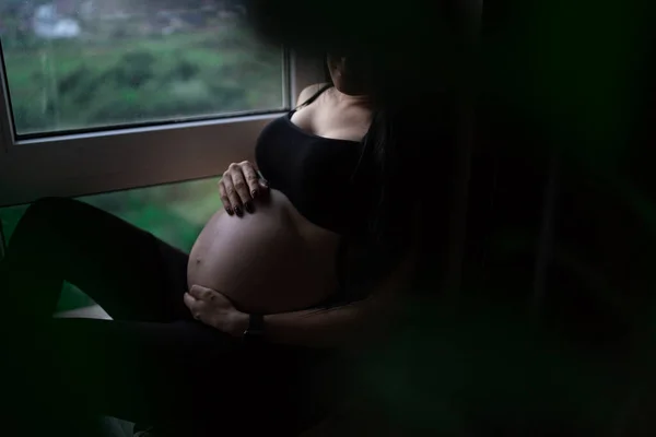 Беременная женщина в чёрной одежде держит руки на животе на тёмном фоне. Беременность, материнство, подготовка и ожидание концепции - крупным планом счастливой беременной женщины с большим животом у окна . — стоковое фото