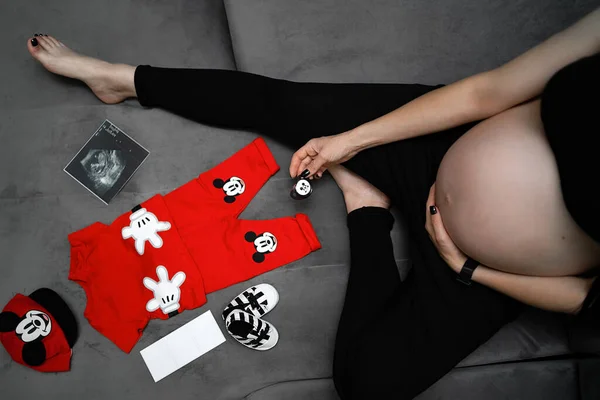 Femme enceinte emballe des vêtements de bébé pour aller à la maternité vue de dessus. Belle jeune femme enceinte assise près de la fenêtre. Elle regarde des vêtements de bébé mignons de rêve et souriant — Photo