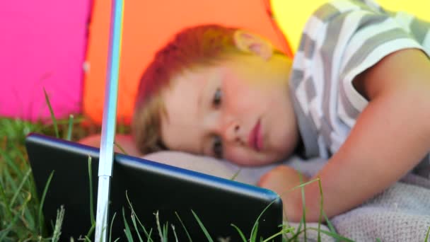 Enfant avec comprimé couché sur de l'herbe verte sous le parasol. Concept vacances d'été . — Video