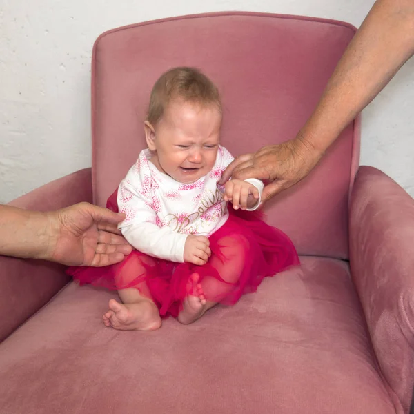 Huilende baby zittend in de stoel. Handen van grootmoeder bedrijf kind zittend in de stoel. Negatieve emoties van het kind. Gelaatsuitdrukking. Portret van aangeklede meisje zittend in de stoel. — Stockfoto