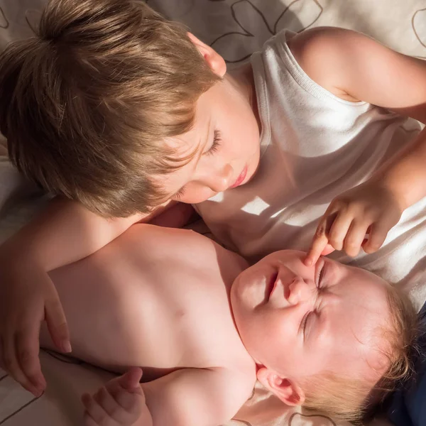 Elder brother is playing with the newborn. Cute boy lovingly playing with his baby sister. The concept of love and friendship between older and younger siblings. Elder brother blonde boy son playing — Stock Image Elder Brother Playing Newborn Cute Boy Lovingly Playing His Baby Royalty Free Stock Photos