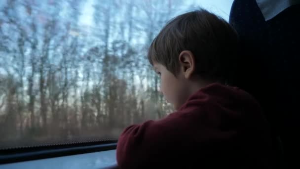 Närbild av en liten nyfiken pojke som tittar ut genom fönstret i tåg. Dess regnar utanför, barn som återspeglar i glas — Stockvideo
