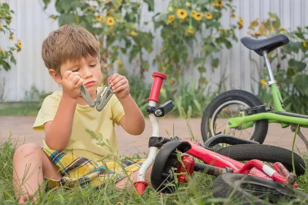 Ребенок открывает мультиинструмент, сидя рядом с велосипедом. Мальчик чинит велосипед. Летние каникулы. Летние лагеря фон. Транспорт и дети. Велосипед на траве. Мальчик играет на улице . — стоковое фото