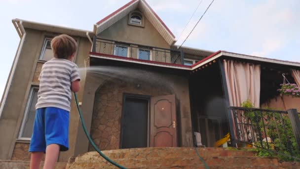 Chico lavando la casa al aire libre. Manguera de agua Actividad exterior de verano para niños. Concepto de infancia feliz. Niño ayudando a los padres a hacer las tareas . — Vídeo de stock