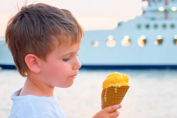 Αγόρι με παγωτό με γιοτ στο παρασκήνιο. Γλυκό φόντο φαγητού. Παιδί που τρώει παγωτό στην παραλία με πλοίο στο παρασκήνιο. Γλυκιά ζωή, εννοιολογική. Η γεύση της ζωής. Καλή ιδέα για την παιδική ηλικία — Φωτογραφία Αρχείου