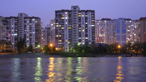 Complexe de bâtiments d'appartements dans la ville du soir près du grand lac, avec des lampadaires se reflétant dans l'eau. Bâtiments au crépuscule près du lac. Bâtiments et lumières dans les appartements le soir . — Video