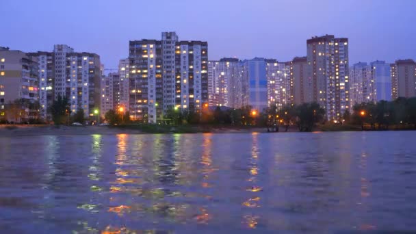 Appartment buildings located near the lake, during sunset with purple sky reflecting in the water. Evening cityscape background. Time lapse. Evening city lights reflecting in the water, river, lake. — Stock Video