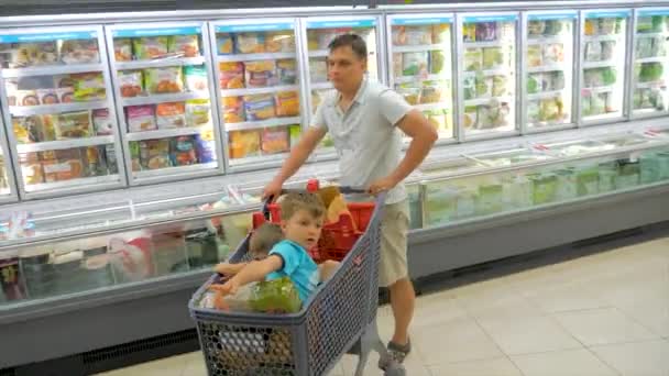 Alışveriş geçmişi. Çocuklar alışveriş sepetinde oturuyor. Babam alışveriş arabasında oturan iki çocuk ile süpermarket etrafında hareket. Süpermarkette yiyecek ve içecek satın almak. Süpermarket ürün seçimi — Stok video