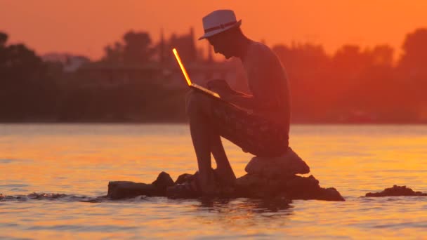 Hombre con portátil en el mar. Hombre con gadget sentado en la piedra en el océano, mar. Hombre trabajando en el portátil con puesta de sol, salida del sol en el fondo. Formación profesional. Ganar dinero en línea — Vídeos de Stock