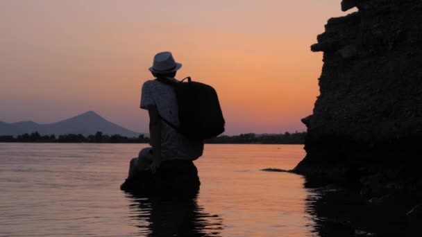 Turista tomando un descanso en la piedra en el mar. Hipster sentado en la roca en el mar en el crepúsculo. Hombre en sombrero con mochila mirando el atardecer en la orilla del mar. Escena no urbana — Vídeo de stock