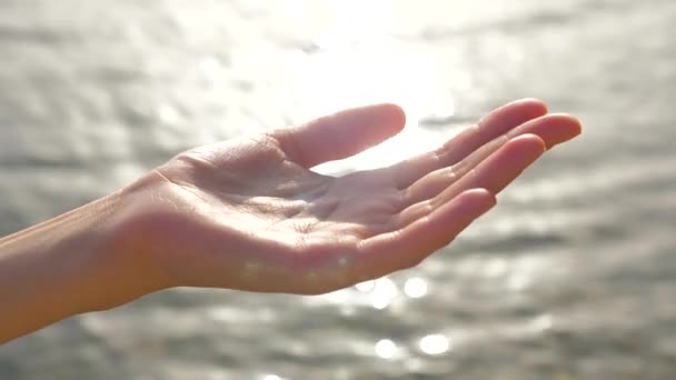 La mano ventosa hembra atrapa las gotas de agua que caen sobre el fondo del paisaje marino. El sol brilla en la mano de la mujer, mientras que las gotas de agua que caen en la palma. Mano de mujer jugando con gotas de agua — Vídeo de stock