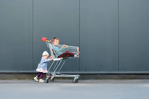 Due hipster alla moda con carrello vicino al supermercato, centro commerciale. Due bambini carini in attesa di genitori all'aperto. Felice infanzia concetto di sfondo. Bambina che gioca con suo fratello — Foto Stock