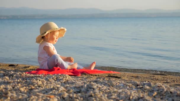 Krásná holčička, která sedí na pobřeží. Malý turista sedí na červeném ručníku a dívá se na mořskou krajinu. Letní čas nebo opětovné vytvoření pozadí. Koncepce rodinných letovisek. Prostředí podporující podporu. Podzimní dovolená — Stock video