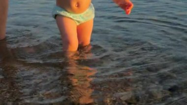 Annem sahilde çocukların ilk adımlarını atmaya yardımcı olur. Bebek deniz suyunda ilk adımları atıyor. Çocuğun bacakları ve anne yürümeye başlayan çocuğa yardım ediyor. Bebeğin başarılı ilk adımları. Pediatri Psikolojisi