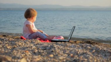 Küçük çocuk deniz kıyısında dizüstü bilgisayarla oturuyor. İş ve tatili birleştiren yeni nesil insanlar. Okul öncesi geçmiş. Eğitimde devrim. Yaratıcılığı geliştirmek. Yeni eğitim sistemi arka planı