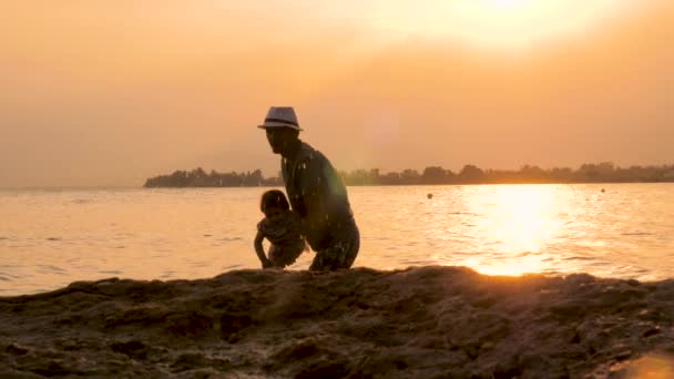 Fiatal apa és a gyerek szórakozik naplementekor a strandon, sziluettek az apa játszik a gyermek a tengerben. apja dobott gyermek fel a levegőbe a tengerbe vízcseppek permetezni körül — Stock videók