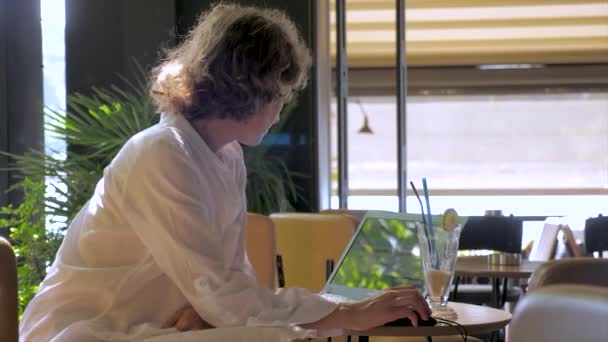 Zavřete portrét mladé ženy v bílých šatech, které sedí u stolu v moderní kavárně a pracují na počítači v netbookem. Čerstvý džus ve skle je u stolu. Série skutečných lidí. — Stock video