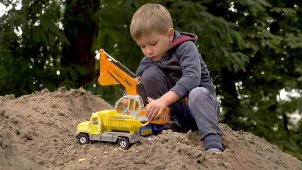 Barnlek grävmaskin och dumpa lastbil i parken, utomhus. En liten byggare lastar sand, jord och jord på en sopbil. Byggkoncept. Pojke leker med leksaker för byggare. Rulla spel bakgrund — Stockvideo