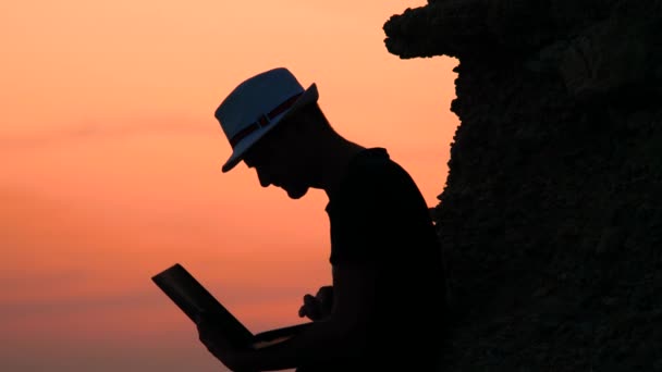 Attractive young man with laptop sitting on the rock of orange sunset on sea shore. Happy vacations time concept. Male freelancer hipster with a laptop remote work on the beach on the sunset — ストック動画