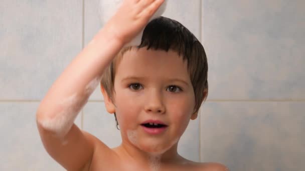 Menino feliz brincando com espuma na sala de banho. Criança a tomar banho. Rapaz alegre a brincar com a água. Cara sorridente de criança. Miúdo a esbanjar água na casa de banho. Actividades internas . — Vídeo de Stock