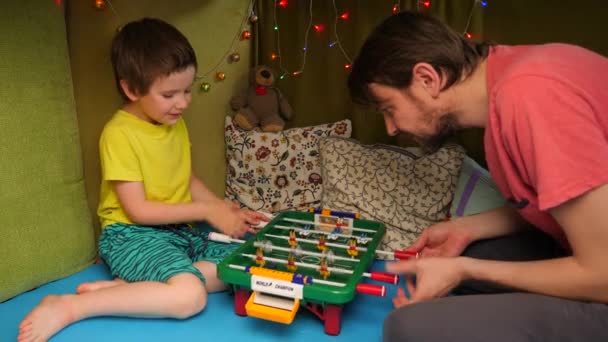 Padre e hijo pasan tiempo juntos en interiores jugando al futbolín. Juego emocional para pasatiempo familiar. Un chico dispuesto a ganar el juego del fútbol de mesa. Cuarentena, entretenimiento en casa — Vídeo de stock