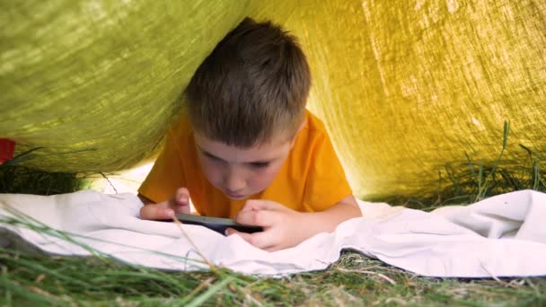 Ребенок читает электронную книгу на открытом воздухе. Парень с гаджетом в парке, в саду. Парень в кепке играет в игру на смартфоне. Обучение, образование онлайн. Дошкольник проводит время на свежем воздухе. Педиатрия, психология — стоковое видео